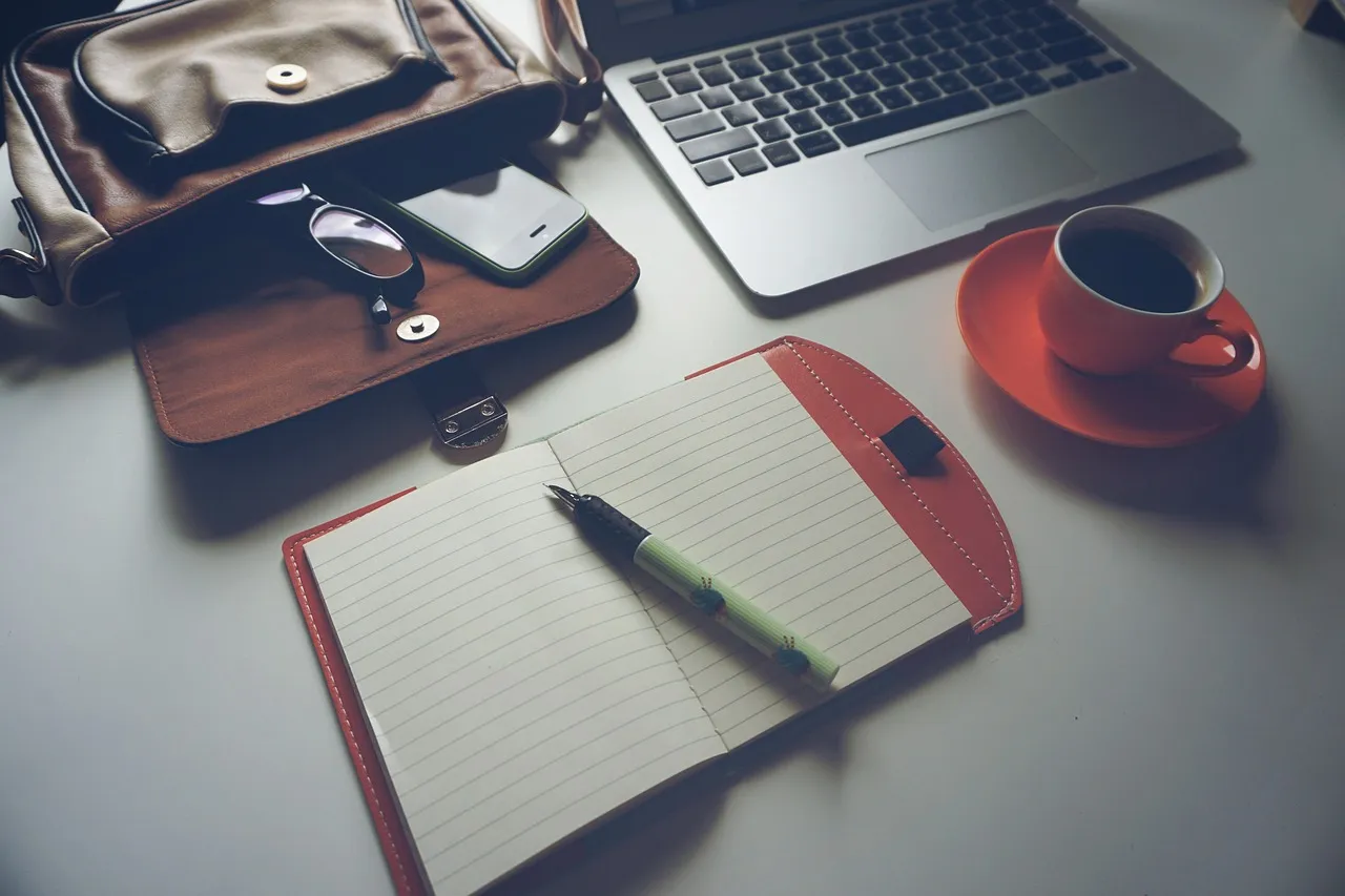 Photo showing laptop, coffee, notebook and pen.